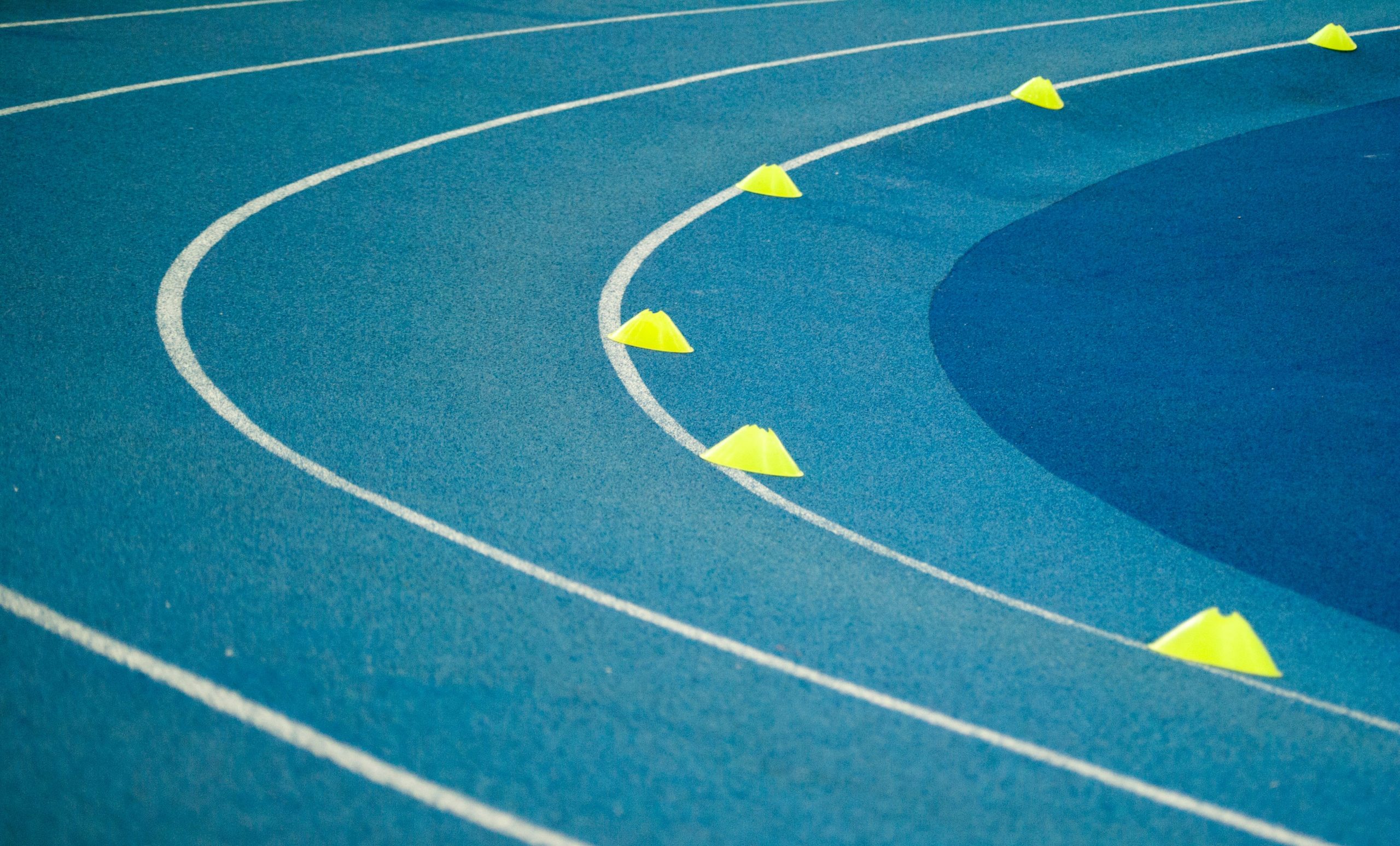 Photograph of a blue running track with white lane markings and yellow traffic cones positioned along a curve