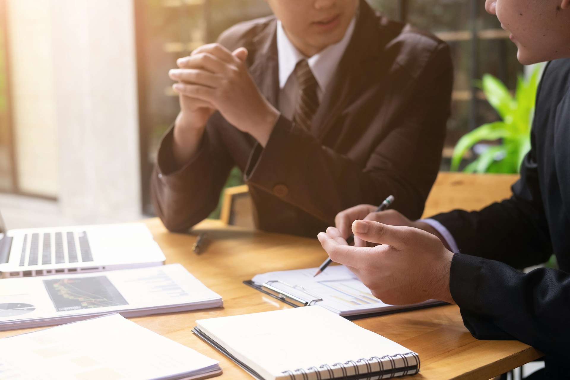 A close-up, cropped shot shows two professionals in dark suits discussing business strategy over a wooden desk