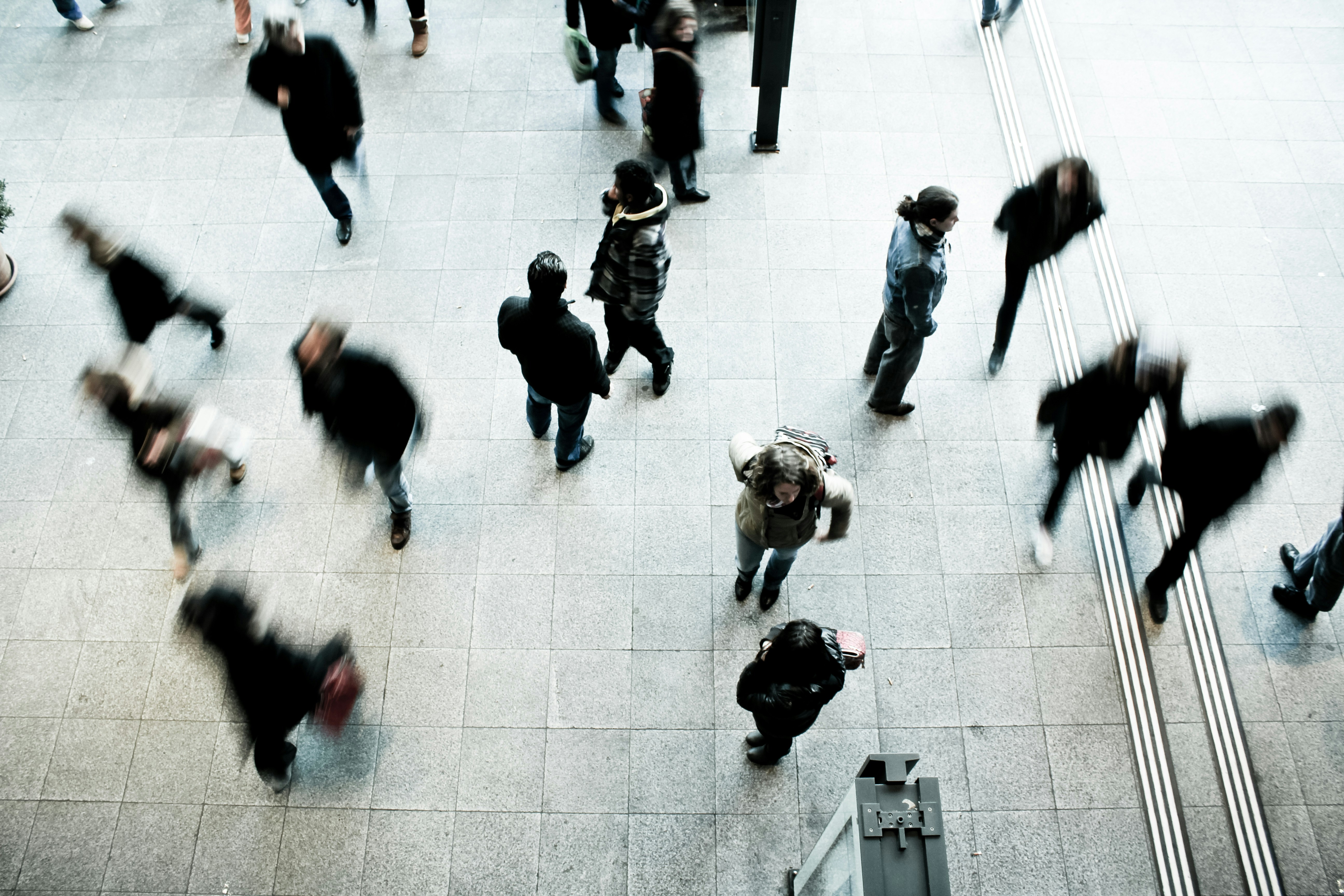 An overhead, high-angle shot captures multiple people walking across a light grey tiled concourse