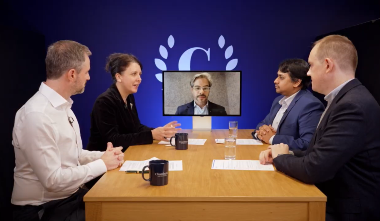 A panel discussion takes place around a light wood conference table against a solid blue background featuring a white "C" logo inside a laurel wreath