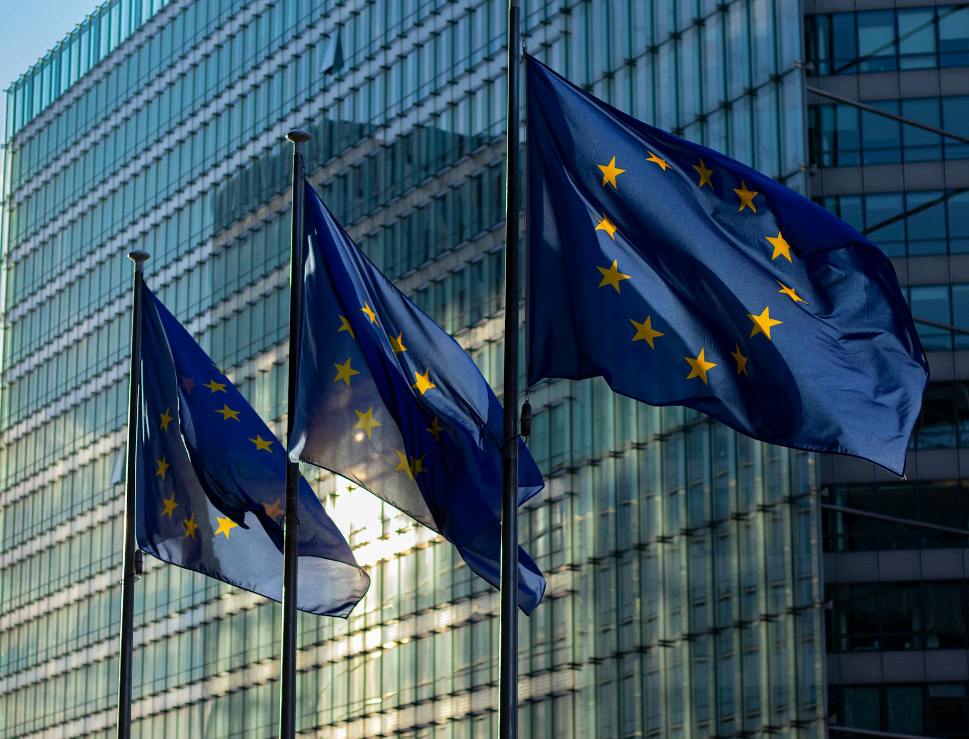 Three European Union flags waving on flagpoles in front of a modern glass office building