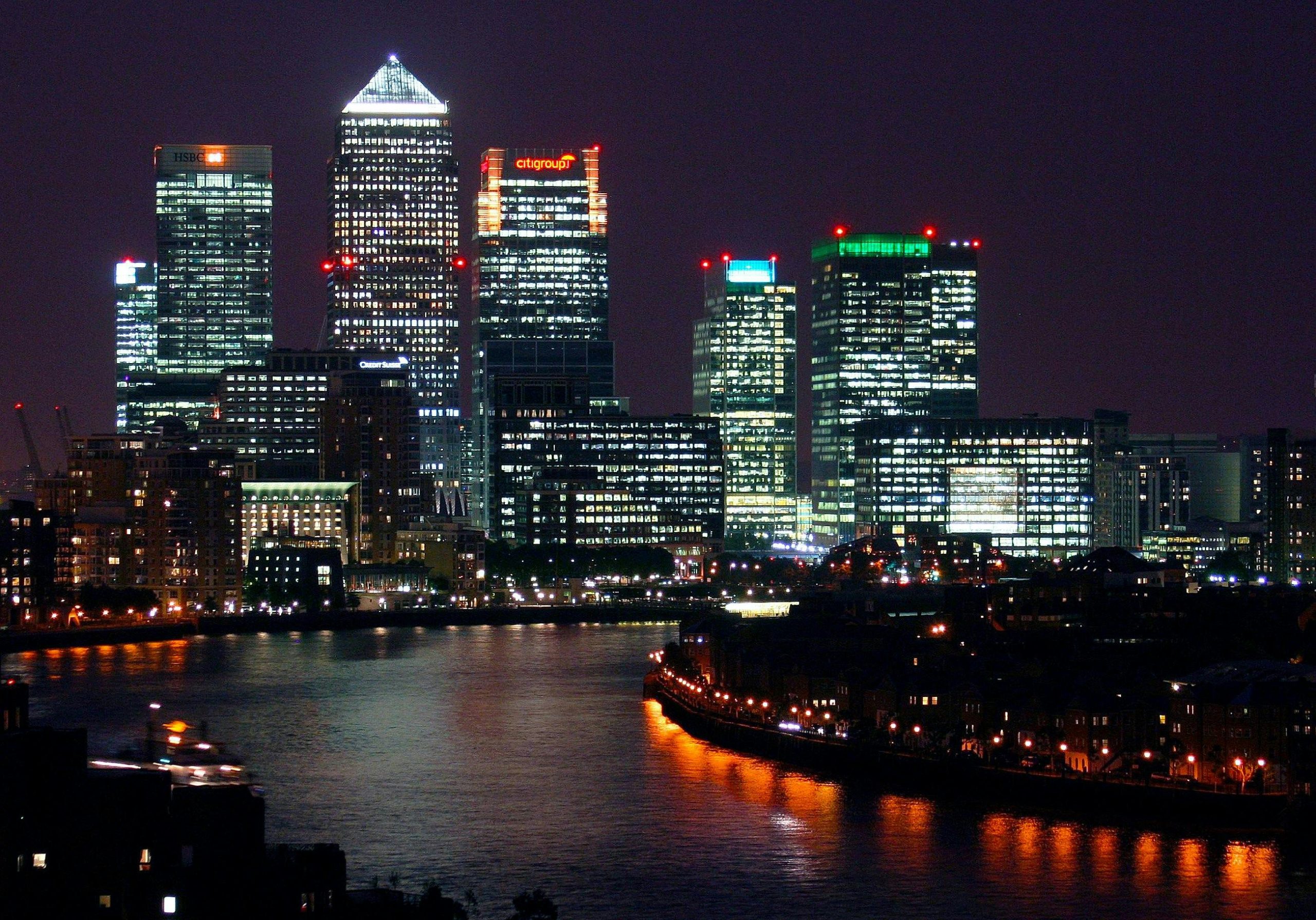Illuminated skyline of Canary Wharf in London at night, with its modern skyscrapers reflecting off the River Thames