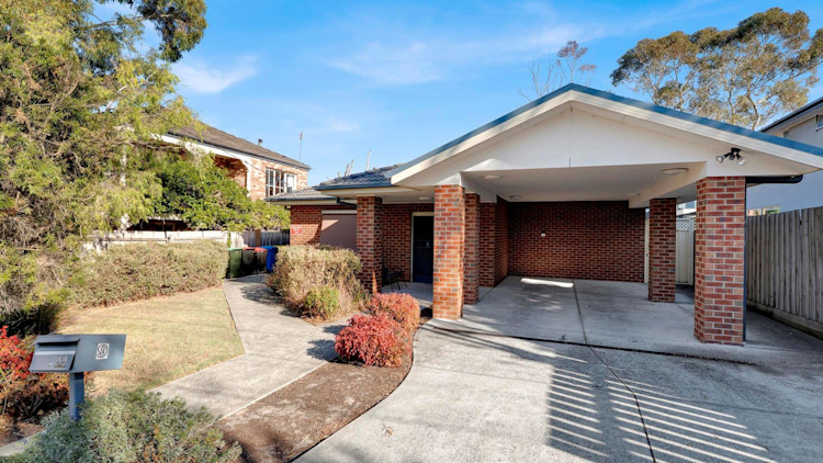 Exterior view of a brick residential home with a covered carport, concrete driveway, landscaped garden, and a pathway leading to the front door, under a clear blue sky.