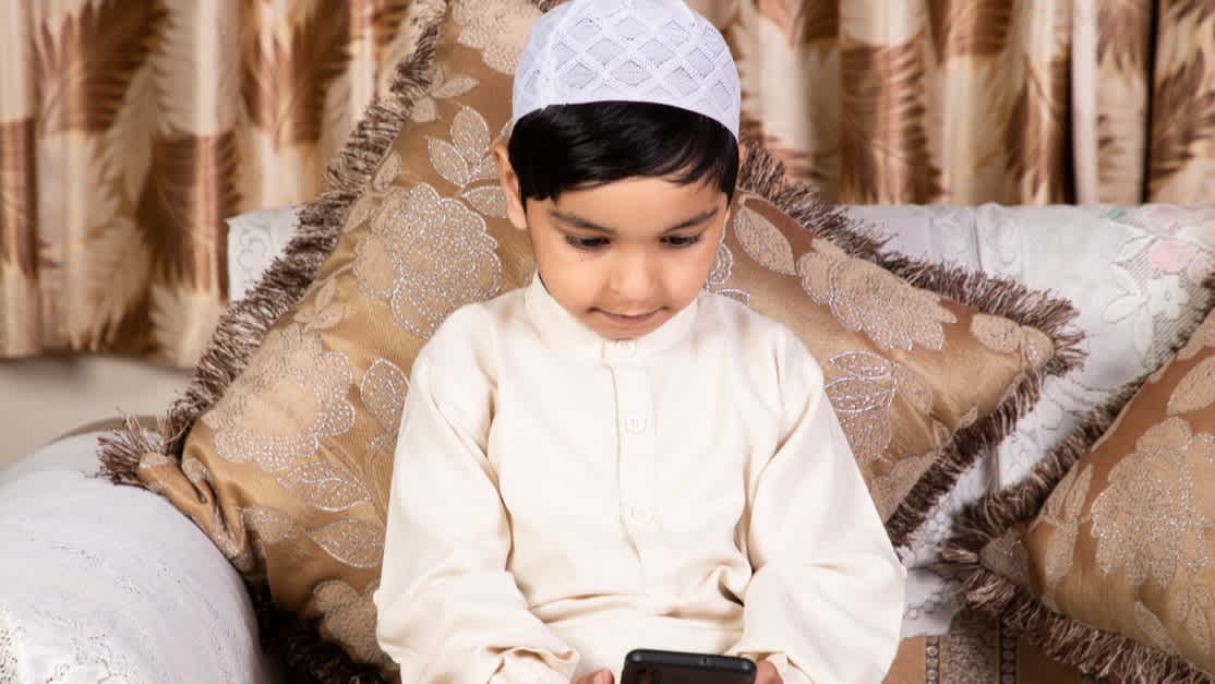 Muslim boy sitting on sofa with mobile phone at home. He is wearing all white.