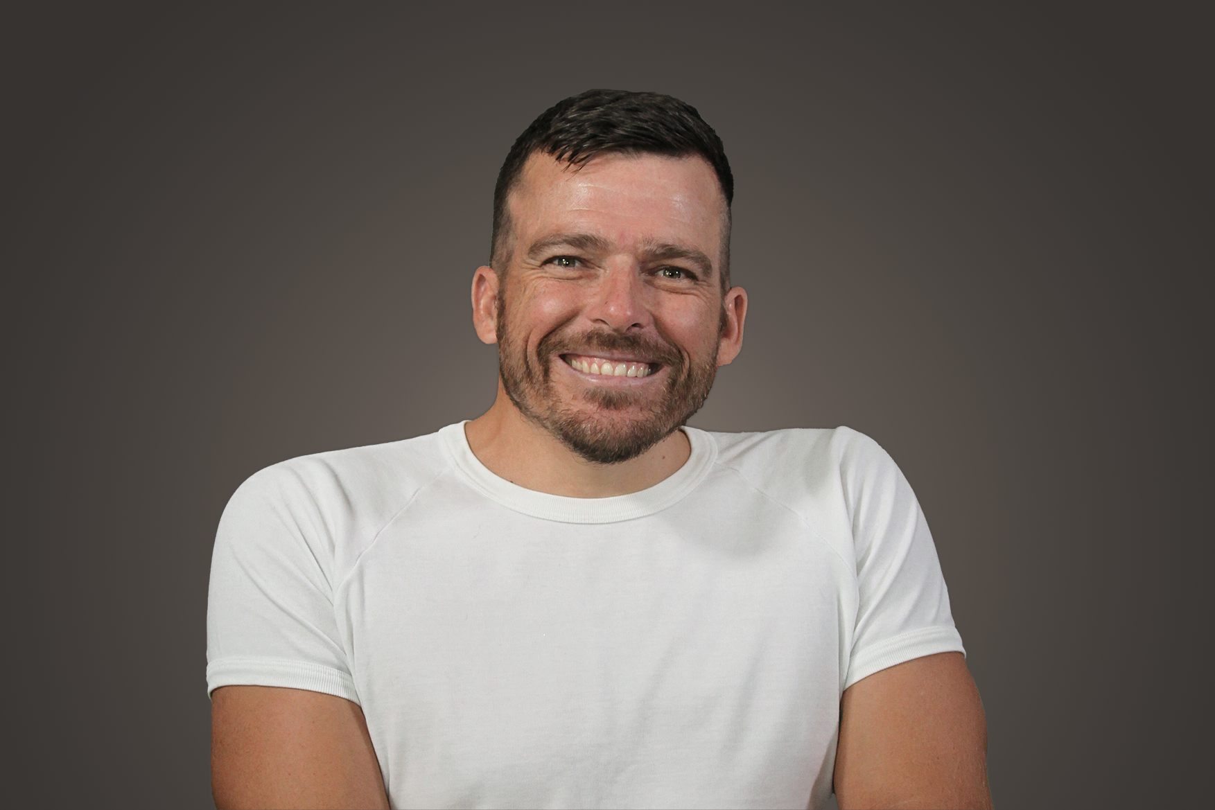 Kurt Fearnley is wearing a white t-shirt and smiling into the camera. He has facial hair and has a dark background.