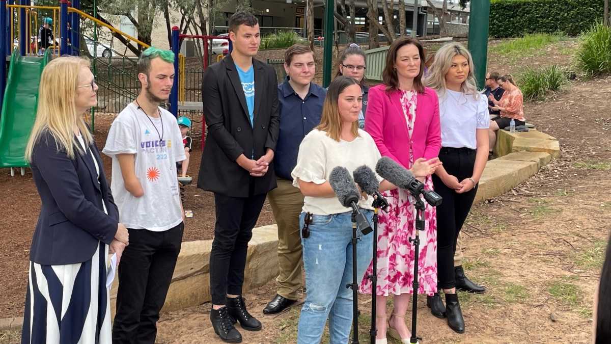 A young woman wearing a yellow shirt and jeans talking into microphones. Behind her is Leanne Linard MP and a group of young people.