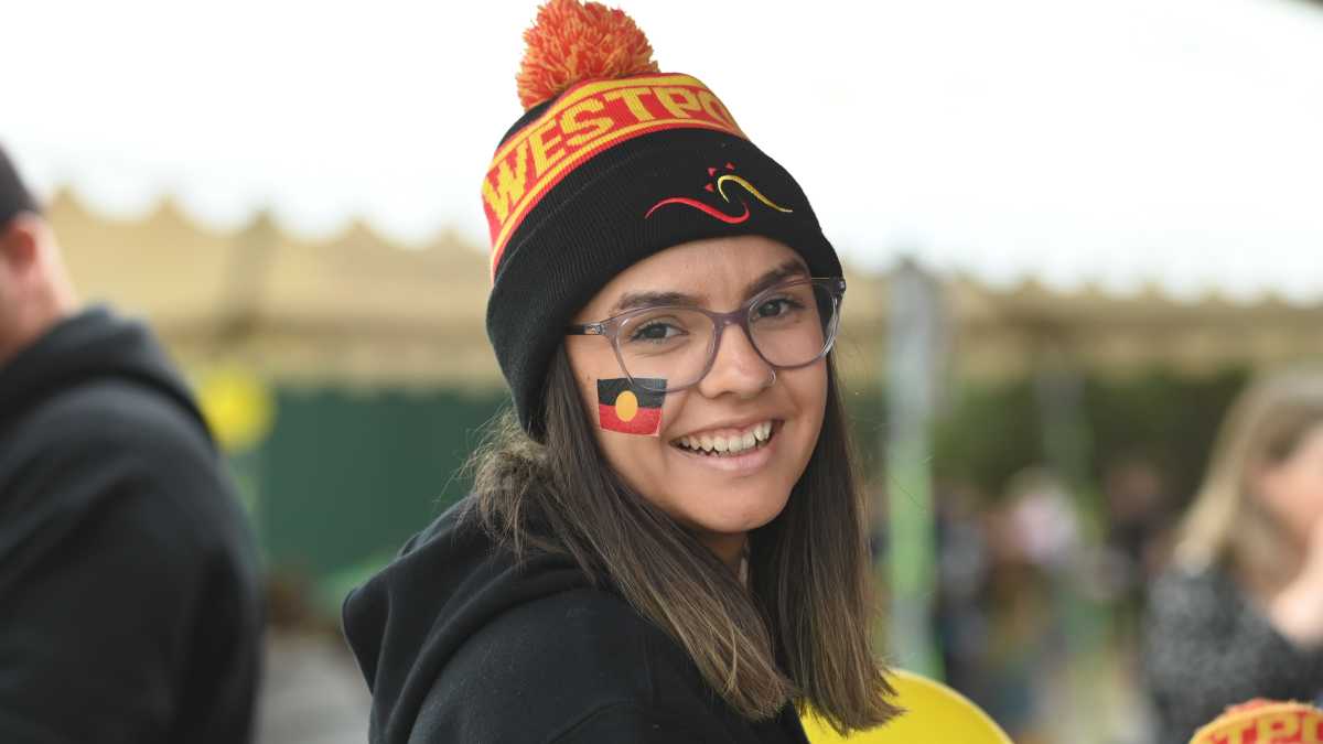 A girl wearing, glasses, a black jumper, and a black, red and yellow beanie, smiling at the camera. On her face is an Aboriginal flag