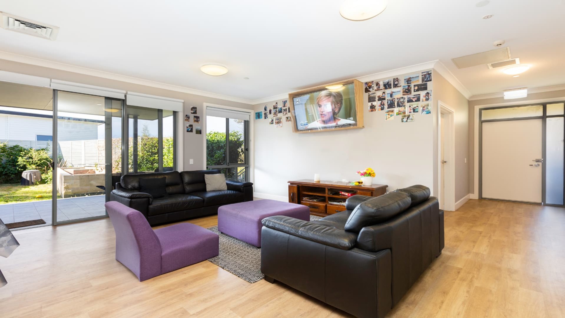 Spacious loungeroom with hard wood floors, a mix of purple and black lounge and chairs, opening out onto the undercover patio.  Wall mounted Television.
