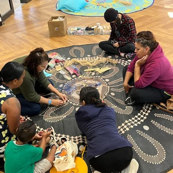 A group of six people are sitting on a black rug with Aboriginal Artwork on it. The group is weaving with coloured thread.