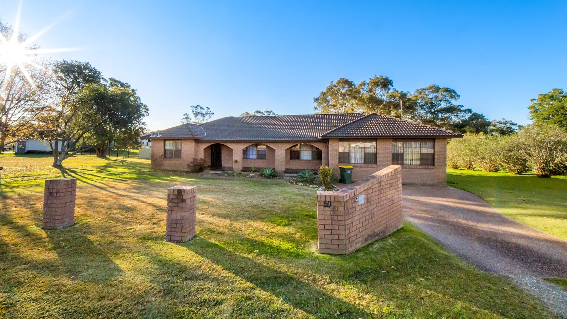 Single-storey brick home with tiled roof, wide front lawn, and brick pillars at driveway entrance.
