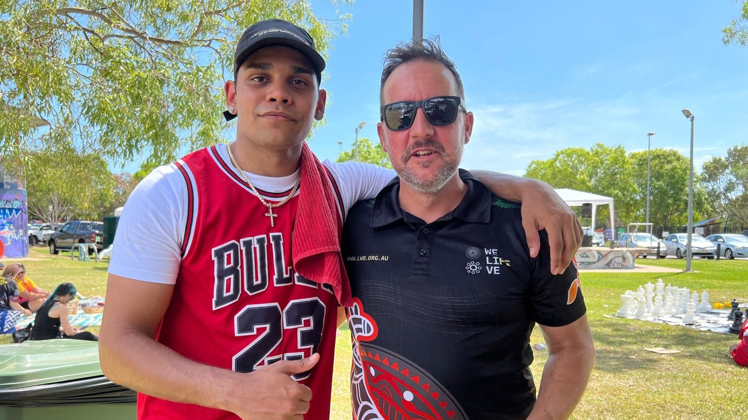 One man in a red singlet stands with his arm around another man in a black polo.