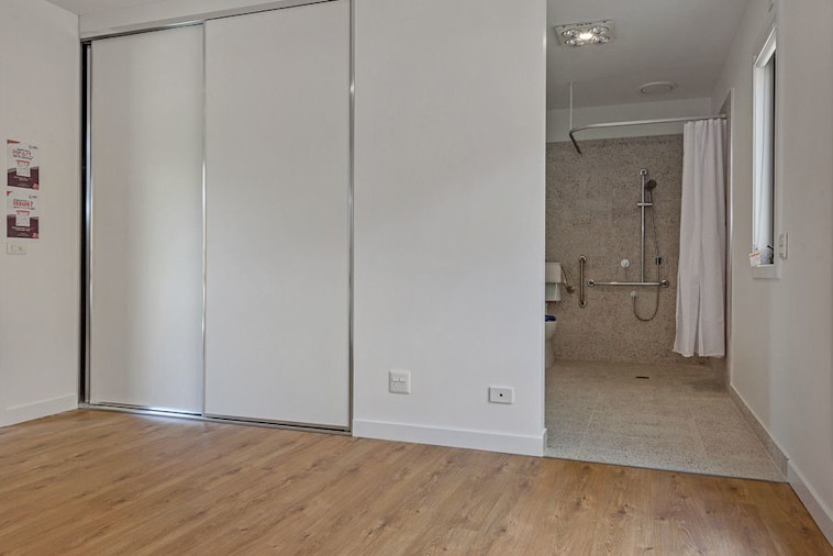 Alternate view of an empty accessible bedroom featuring timber flooring, large sliding wardrobe doors, and direct access to a roll-in ensuite bathroom.