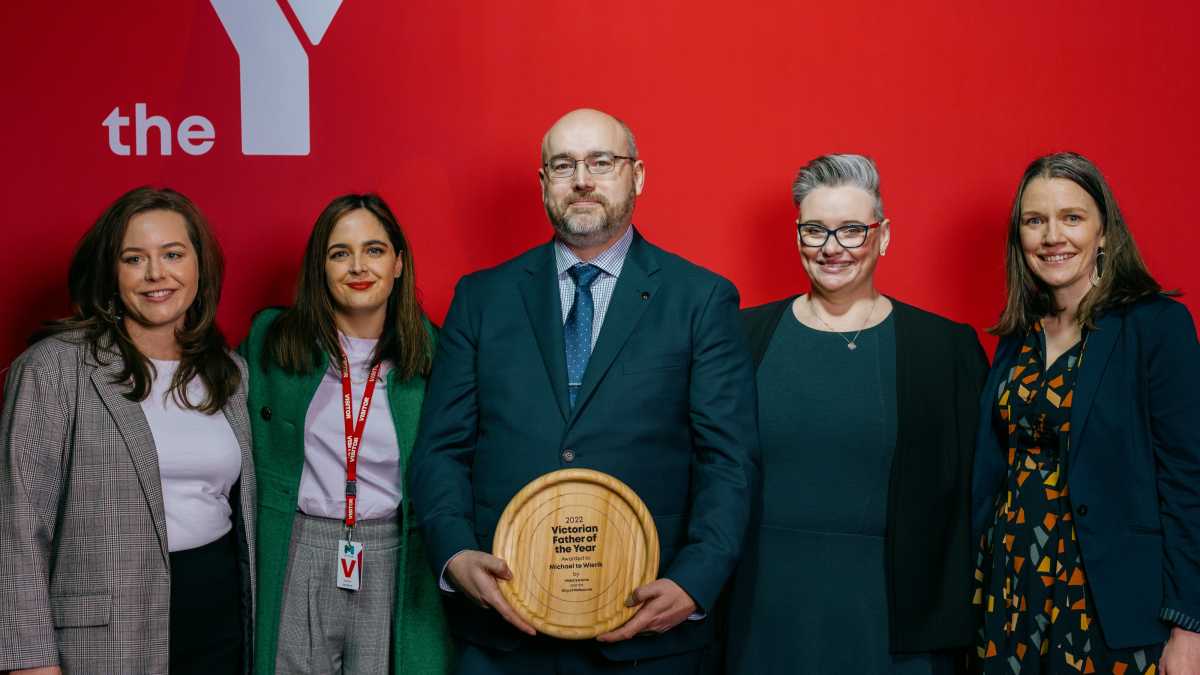 Mike is standing with a group of people in front of a red wall smiling at the camera and holding his award