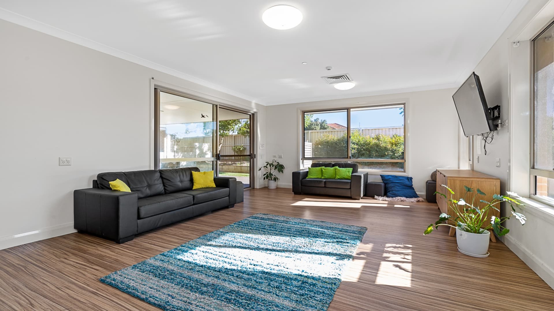 Main loungeroom with two grey couches, blue rug on hard wood flooring, wall mounted television and large windows and sliding door entry to outdoors.