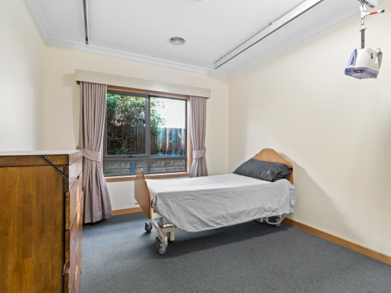 Vacant bedroom features single hospital grade bed, ceiling hoist, grey carpet, chest of drawers, large window with pink curtains.