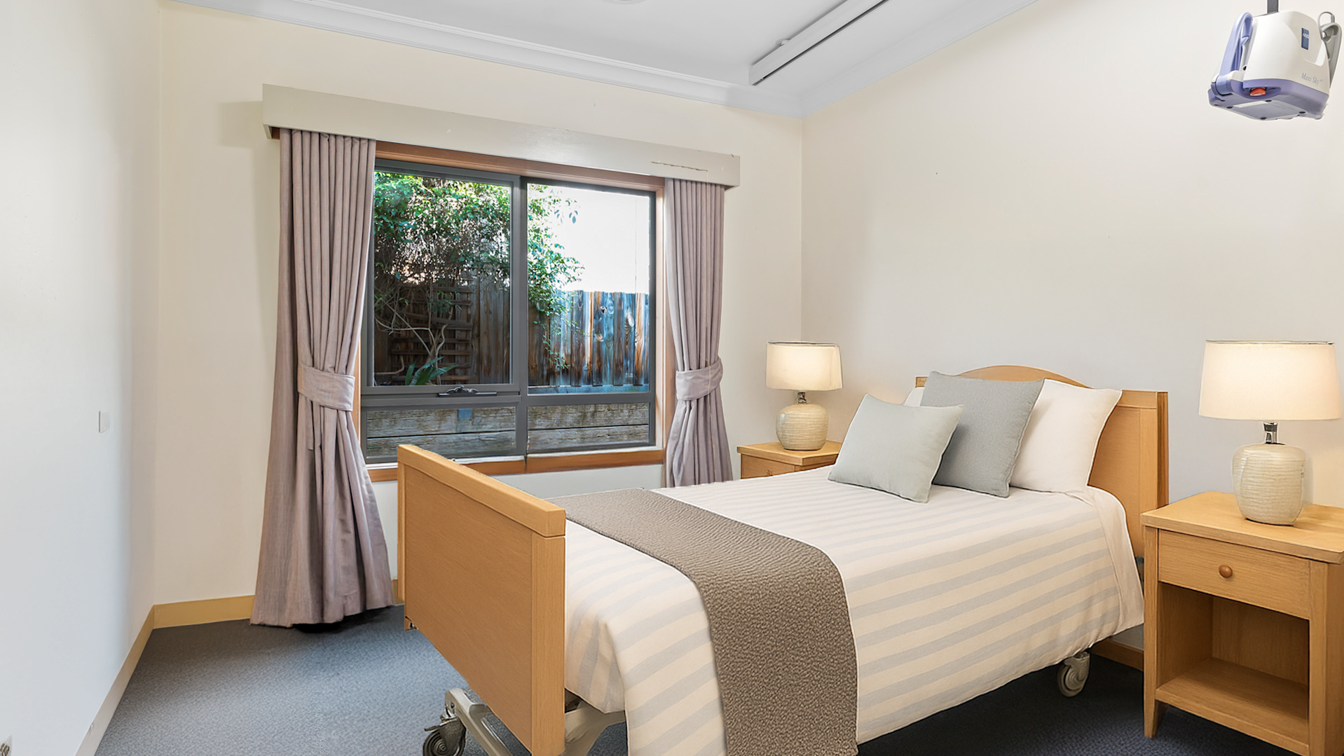 Vacant bedroom features single hospital grade bed, ceiling hoist, grey carpet, chest of drawers, large window with pink curtains.