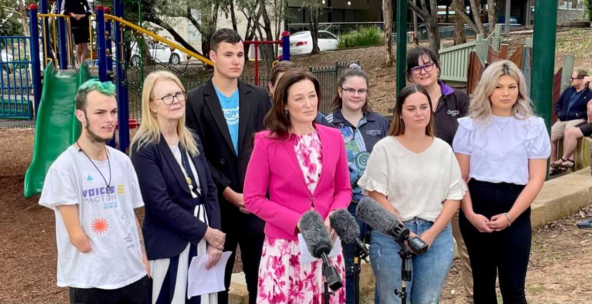Leanne Linard MP wearing a pink floral dress and blazer standing behind microphones with a group of young people.
