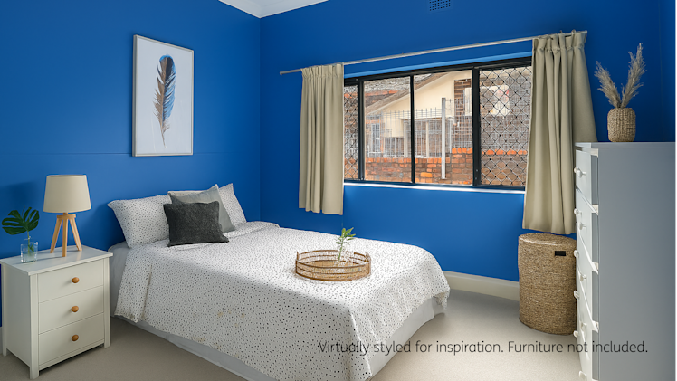 Modern bedroom with deep blue walls, featuring a double bed with white and black spotted bedding, a white bedside table with a lamp and plant, a rattan laundry basket, a white dresser, and beige curtains framing a large window with security mesh.