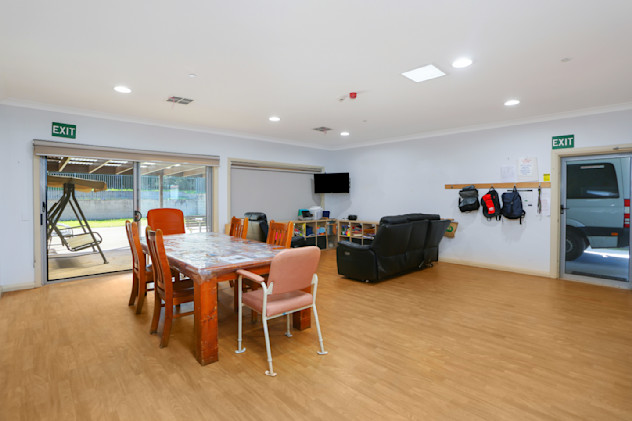 Open dining and lounge area with wooden dining table, mixed chairs, black sofas, wall-mounted TV, and sliding doors to outdoor patio.