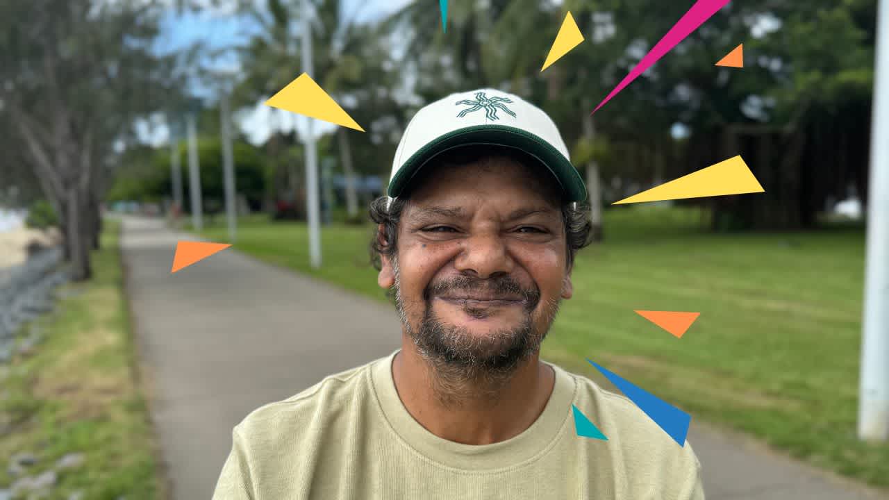 A close up Will's face. He is wearing a cap and tshirt and smiling at the camera, a park can be seen behind him.