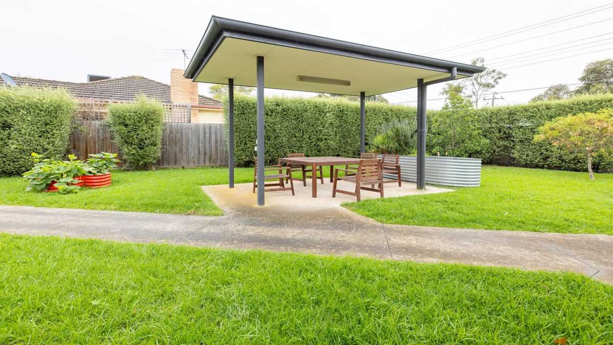 Spacious lawned area with wide concrete walkways, leading to a centrally located covered pergola with table and chairs.