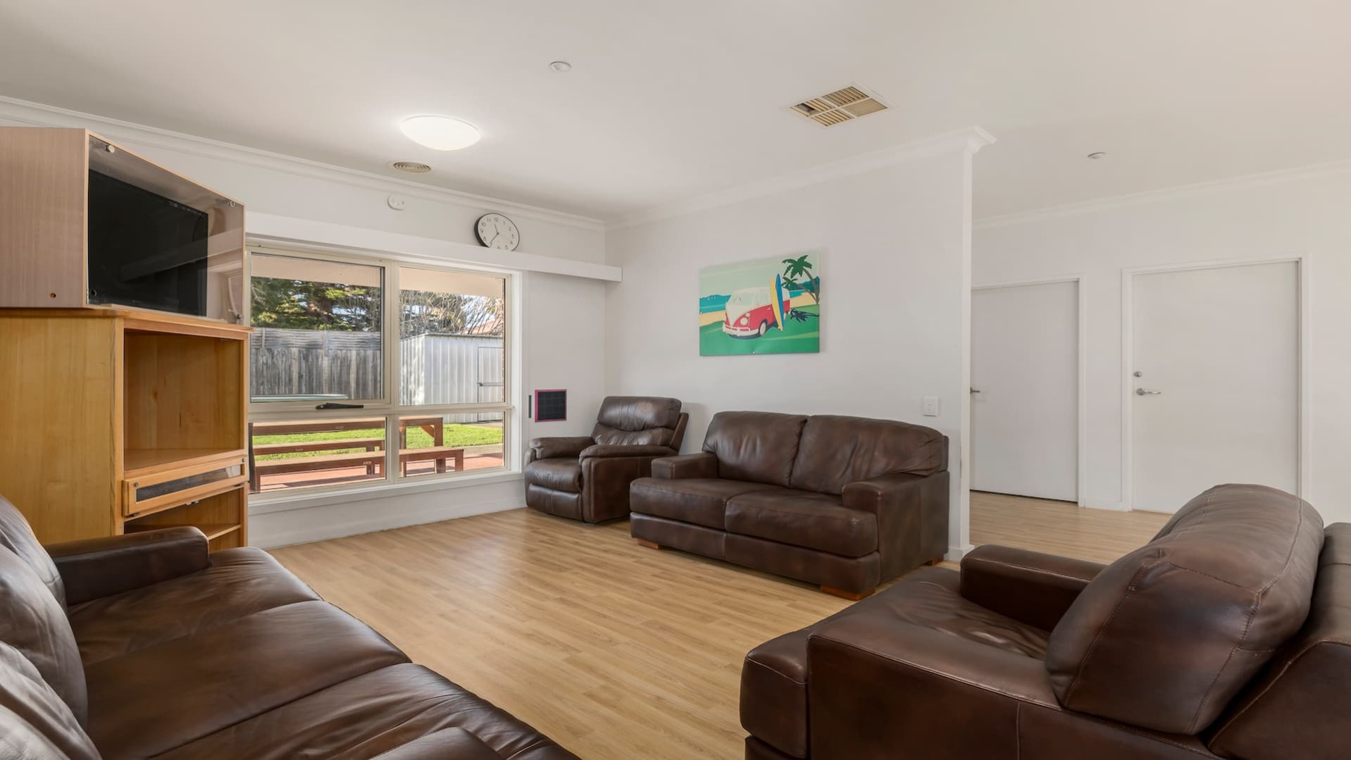 Spacious lounge room with large window, hard wood flooring, two brown leather sofas and two lounge chairs.