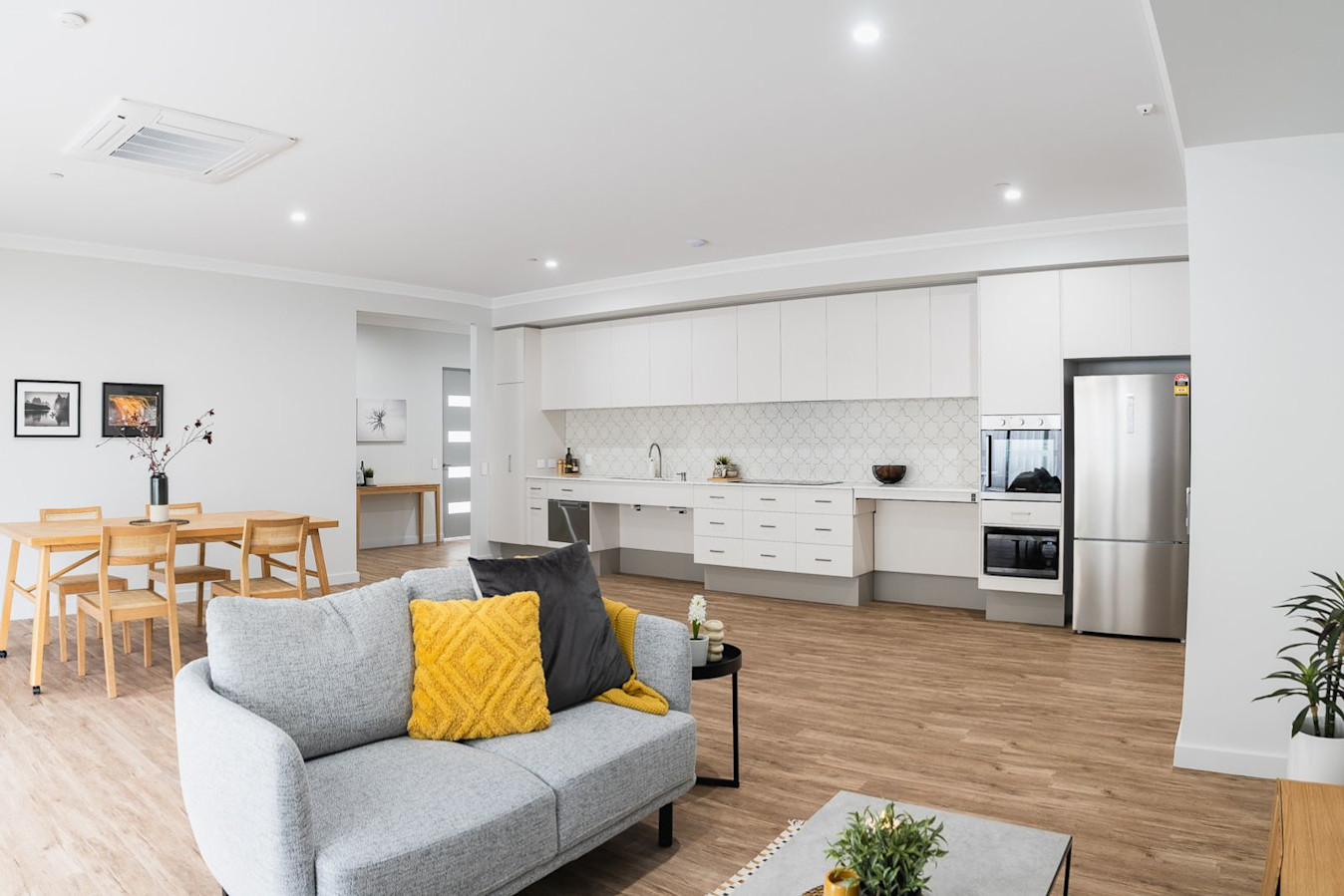 Open-plan living with grey sofa, yellow cushions, wooden dining table, and modern kitchen in background.