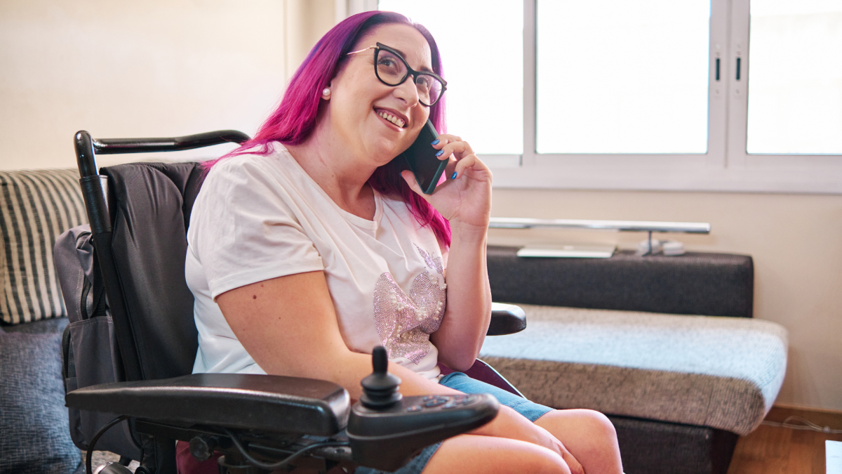 A woman with pink hair wearing glasses and a white t-shirt is sitting in a wheelchair using a mobile phone.