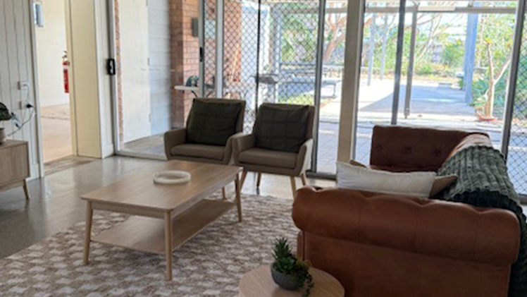 Comfortable living room with a brown leather sofa, armchairs, and coffee table, opening to a tiled outdoor area through glass sliding doors.