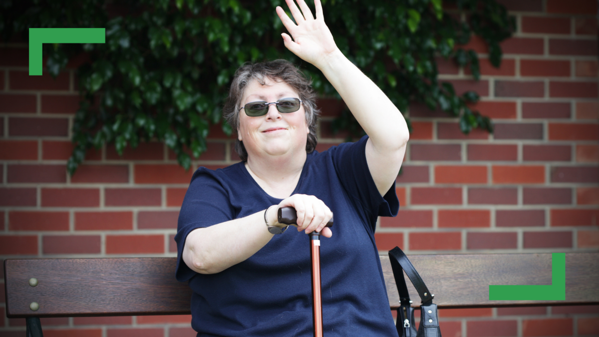 A woman wearing a blue t-shirt and sunglasses is sitting on a bench holding her walking stick and waving at the camera.