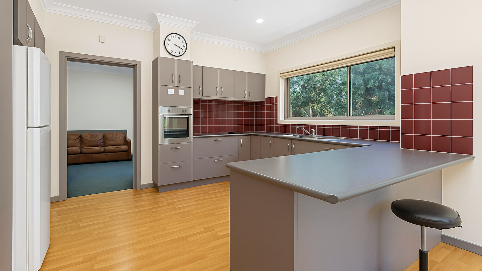 Modern kitchen with brown cabinetry, red tiled backsplash, stainless steel appliances, and a large window overlooking greenery. A lounge area with a leather couch is visible in the adjoining room.