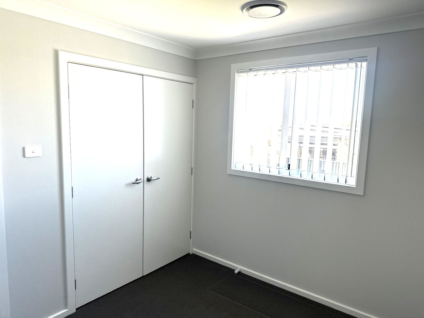 Empty bedroom with grey carpet, white double-door closet, and a window with vertical blinds letting in natural light.