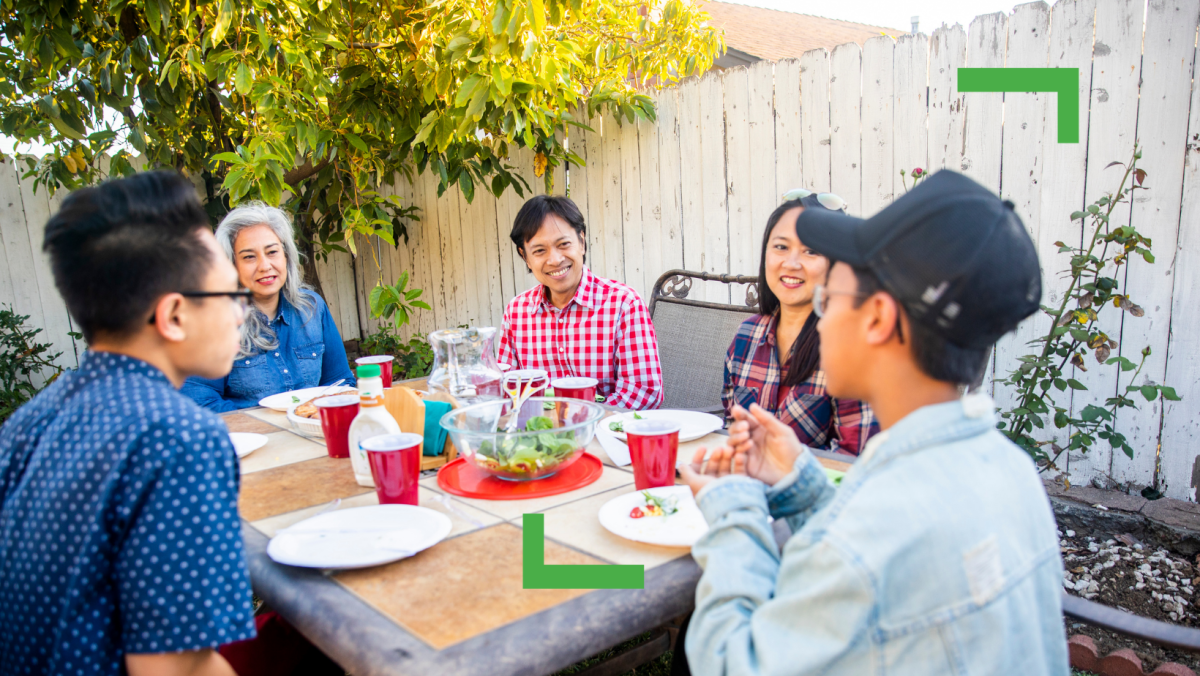 Five people sitting on an outdoor table. Two teenage posts, a father and mother figure. Grandma figure. There is a white fence and tree behind them.