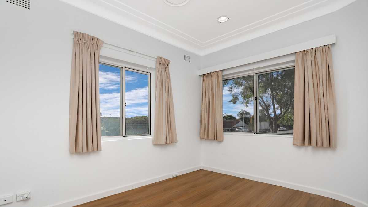 Roomy vacant bedroom with light carpet on the floors and cream curtains on the two windows.