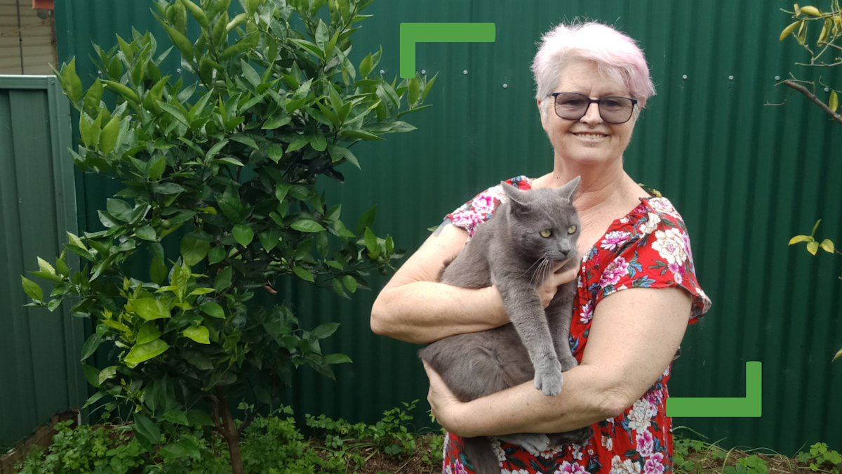 Corrina with her grey pet cat. She is wearing a red flower dress and standing in front of a green shed