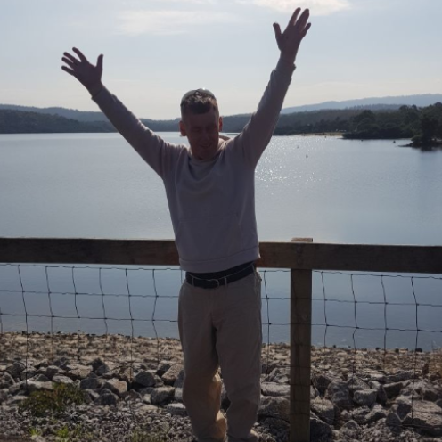 Glen is wearing tan pants and a white jumpber. He stands in front of a fence and a large body of water with his hands in the air.