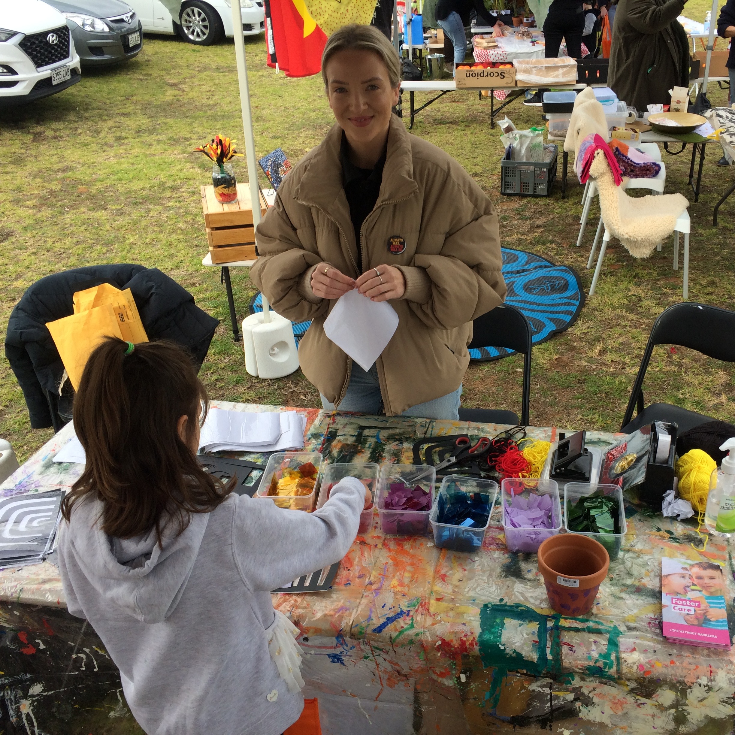 A woman stands behind a table with a craft activity set up. A child stands in front of the table completing the activity.