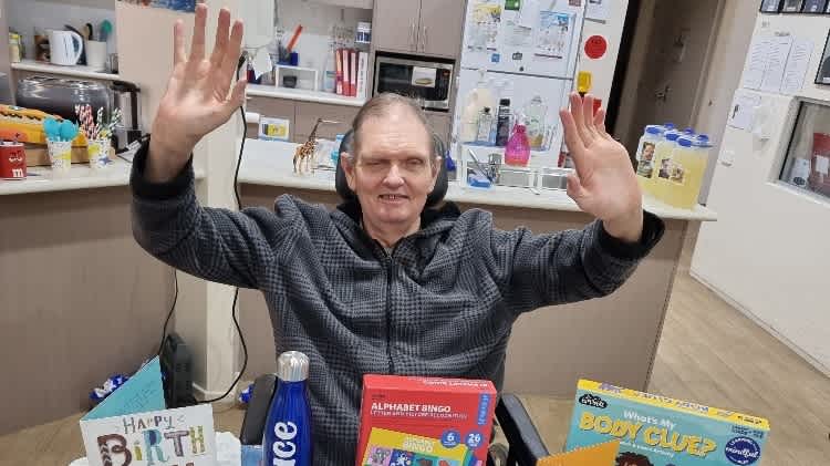 Bruce sits at a table with his hands in the air, smiling at the camera. On the table are birthday cards and gifts.