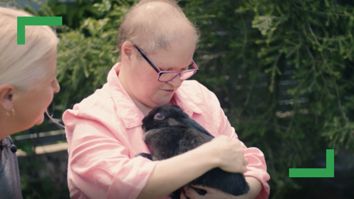 Raelene in a pink shirt and black glasses sitting outdoors holding a black rabbit