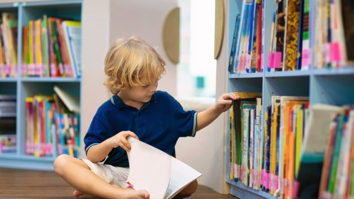 Young blonde boy reading in a library. He is wearing a blue tshirt.
