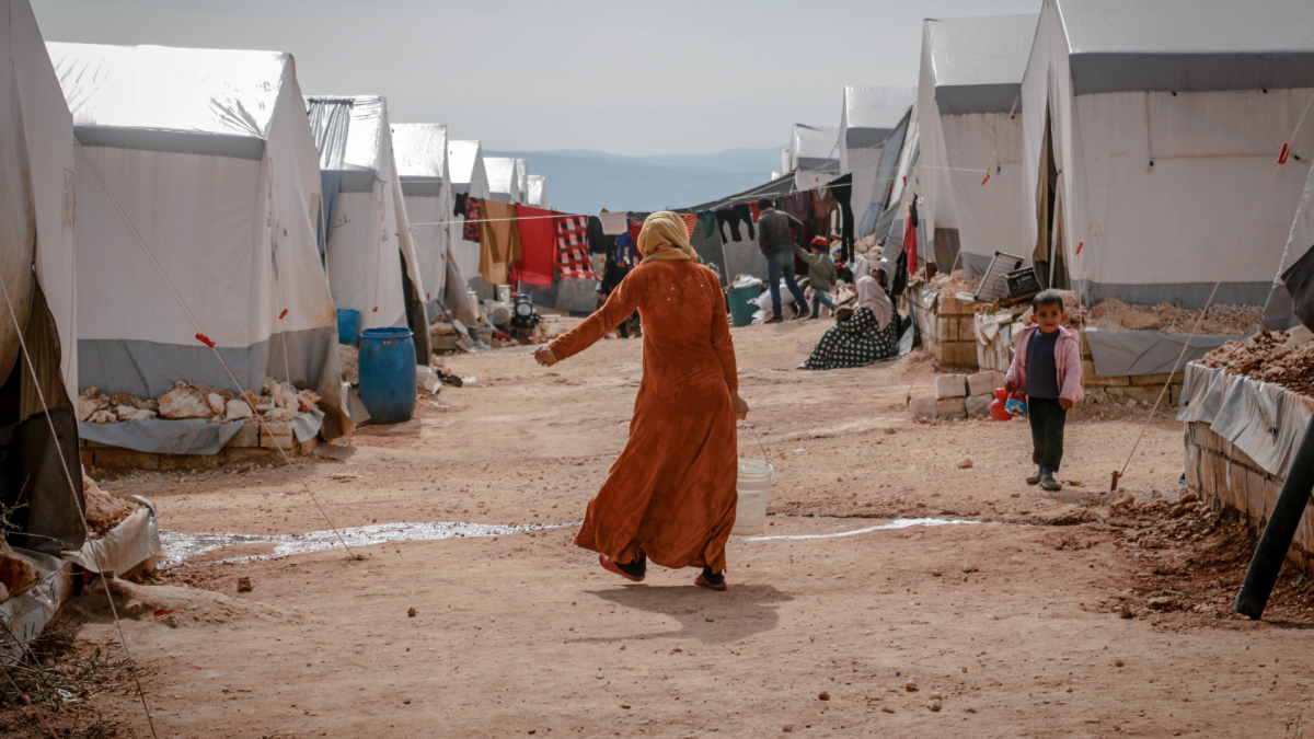 A woman in an orange dress and headscarf walking through a refugee camp holding a bucket.