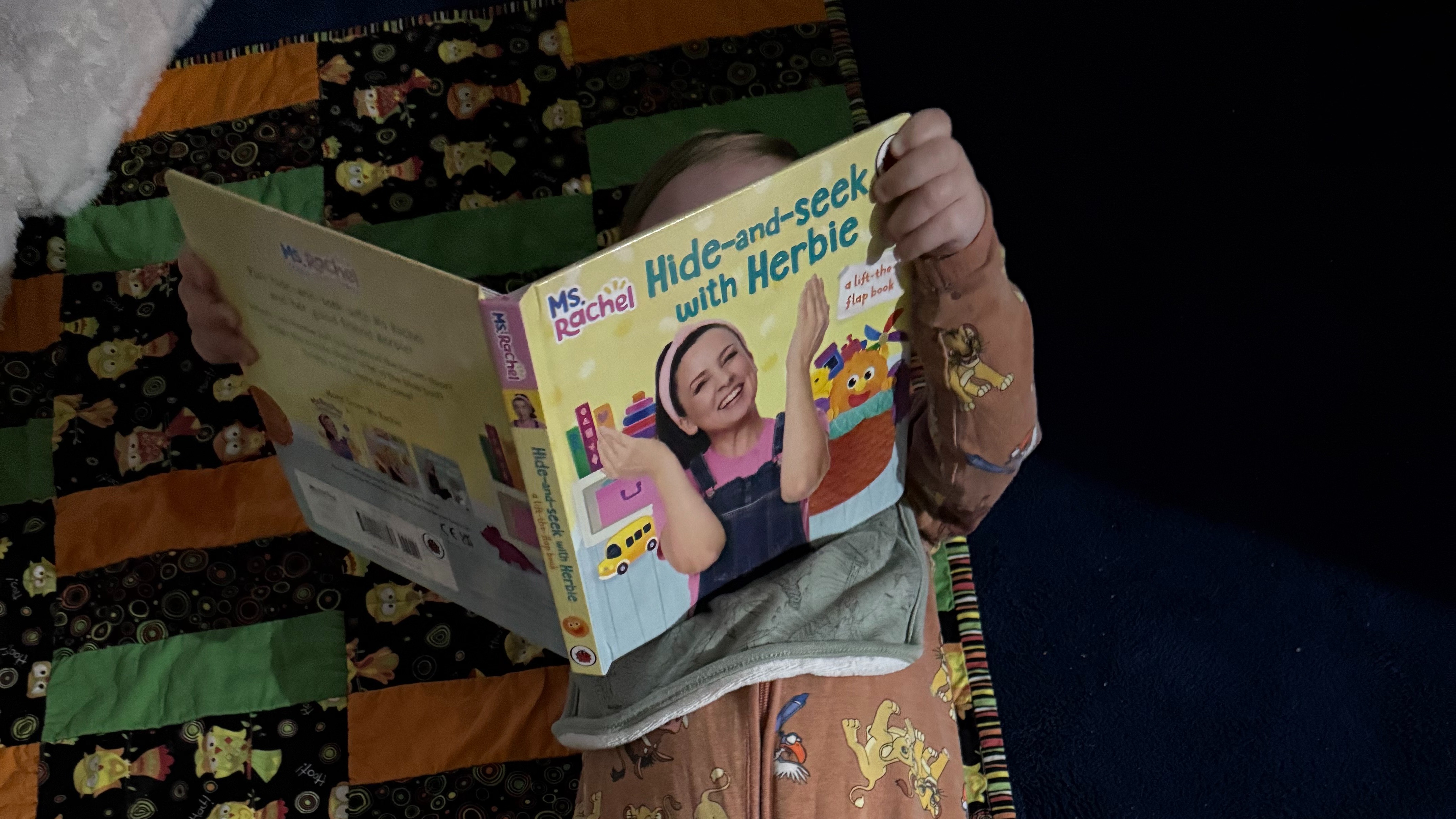 A young child lies on the floor, holding a picture book over their face.