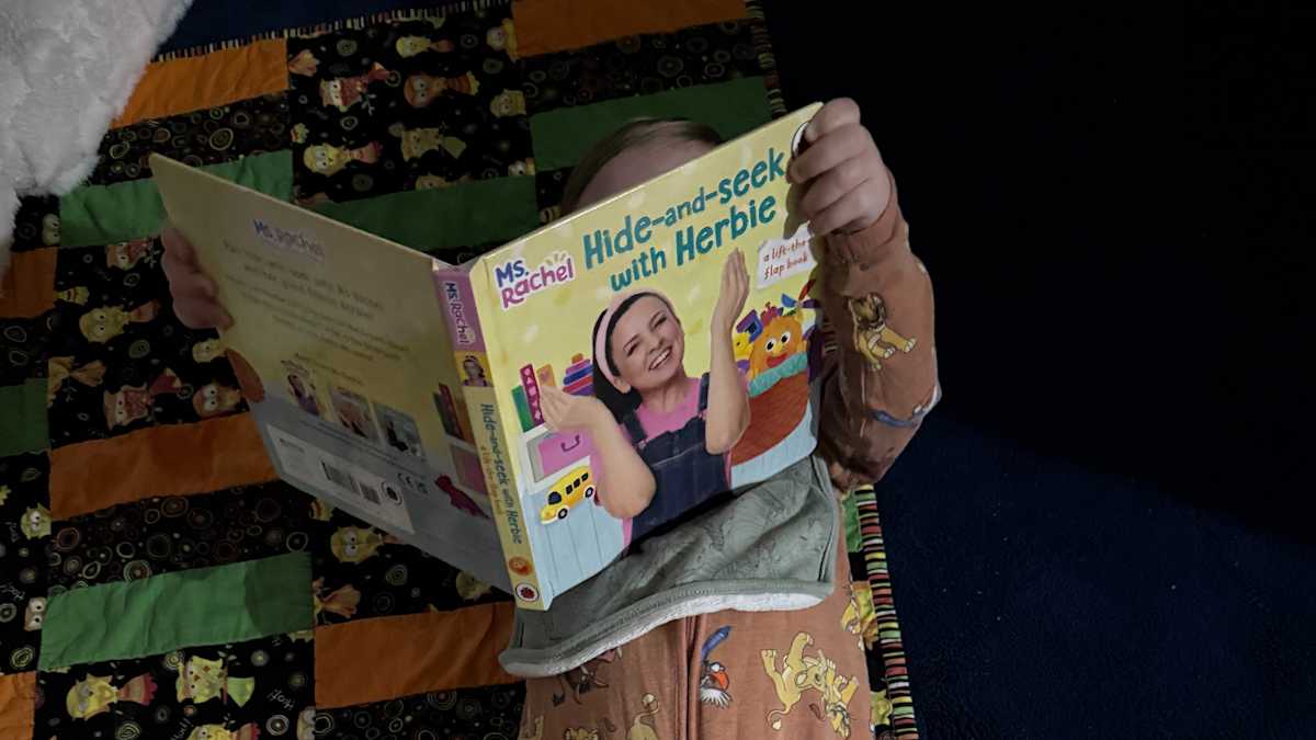 A young child lies on the floor, holding a picture book over their face.