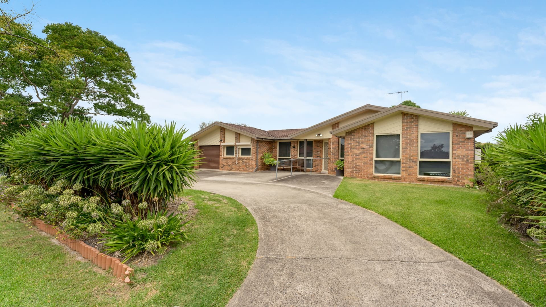 House with a brick facade and a concrete driveway with grass and shrubs on either side leads to the garage and the front door.