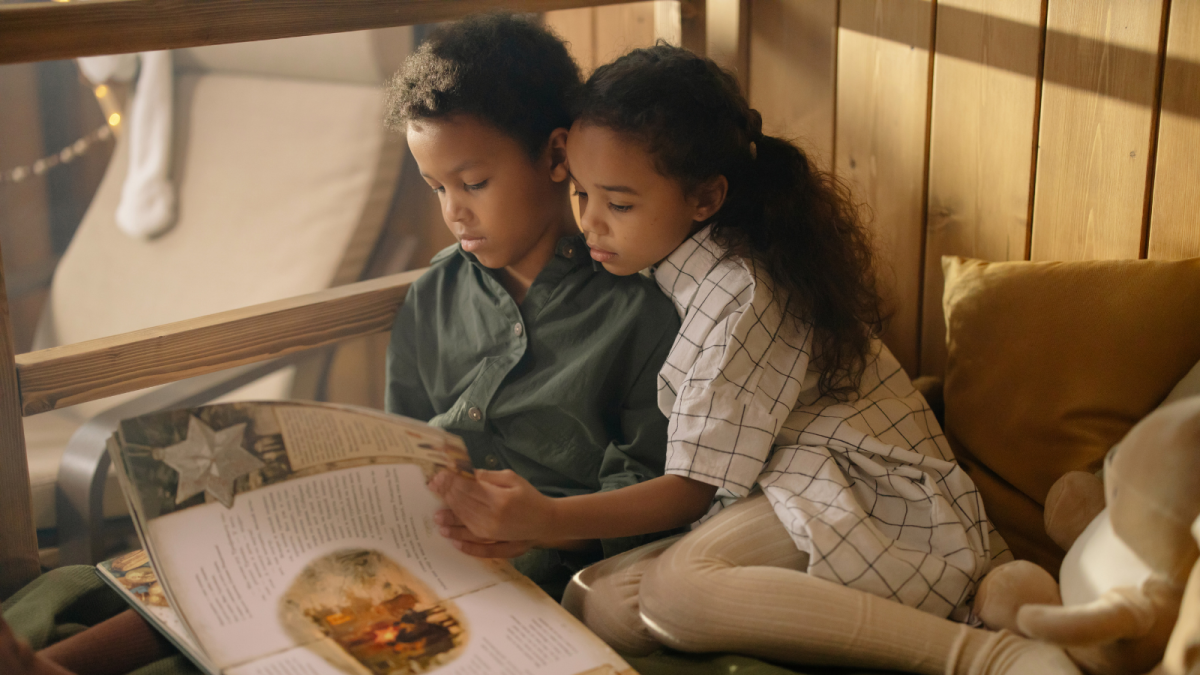A boy in a green shirt with a girl in a white dress and tights are sitting on a bed reading a book.