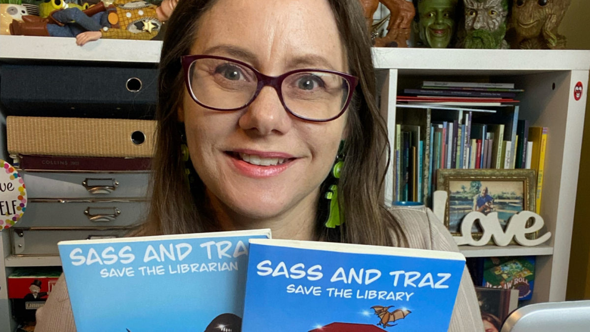 Michelle wearing glasses and sitting in front of a bookshelf holding two copies of her book 'Sass and Traz Save the Library'.