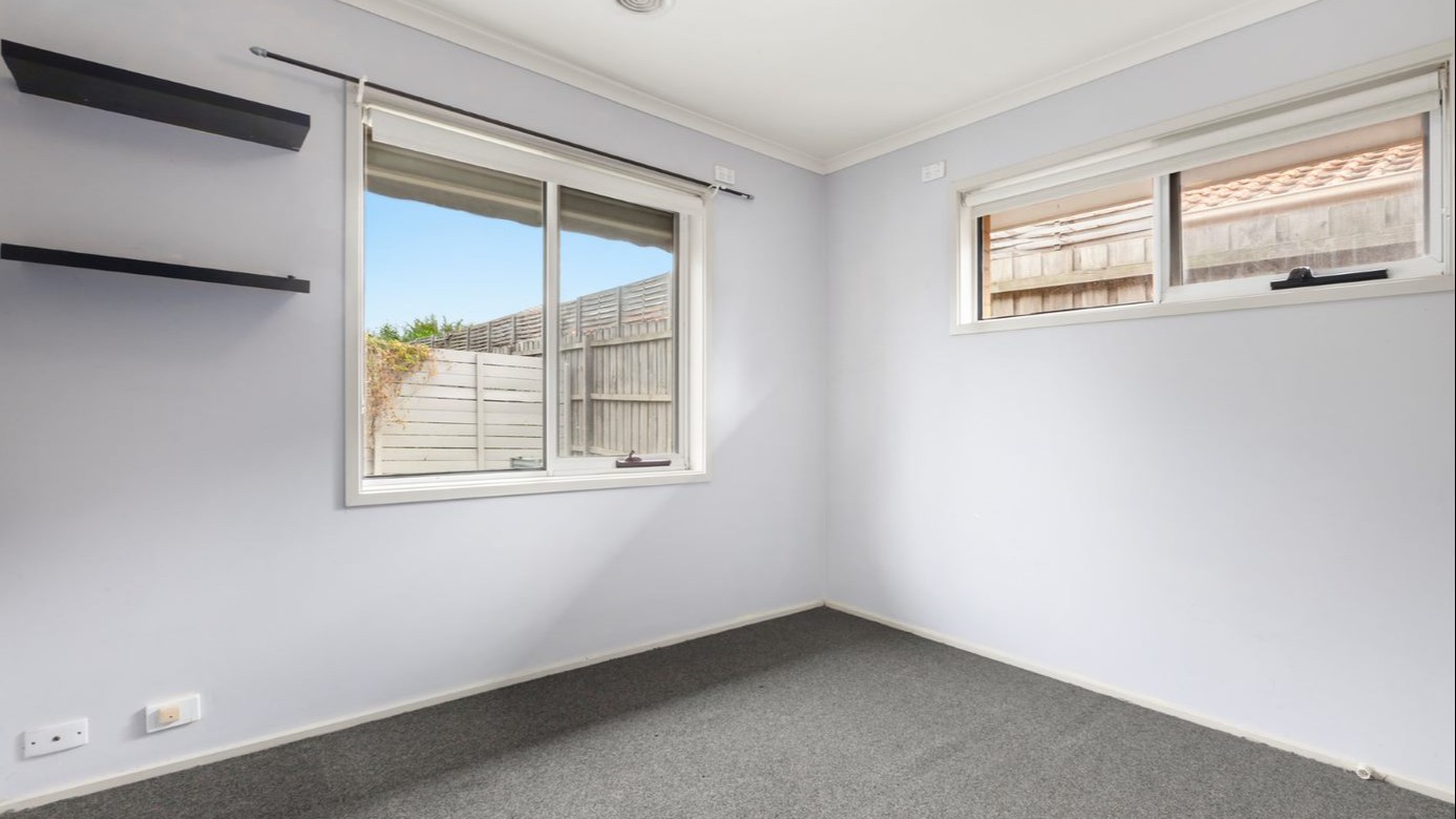 Empty bedroom with dark grey carpet, two windows and two floating shelves on the wall.