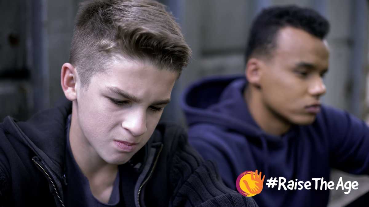 Close-up of two boys looking towards the ground. Raise the age logo and hashtag in the bottom right-hand corner.