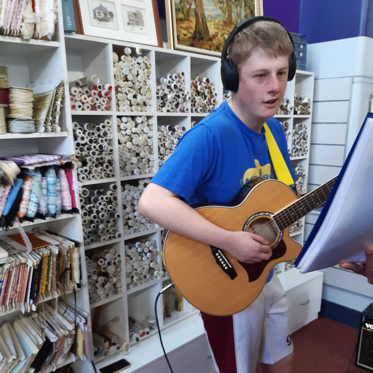 Alex wearing head phones, a blue t-shirt and white shorts standing with a guitar and performing.