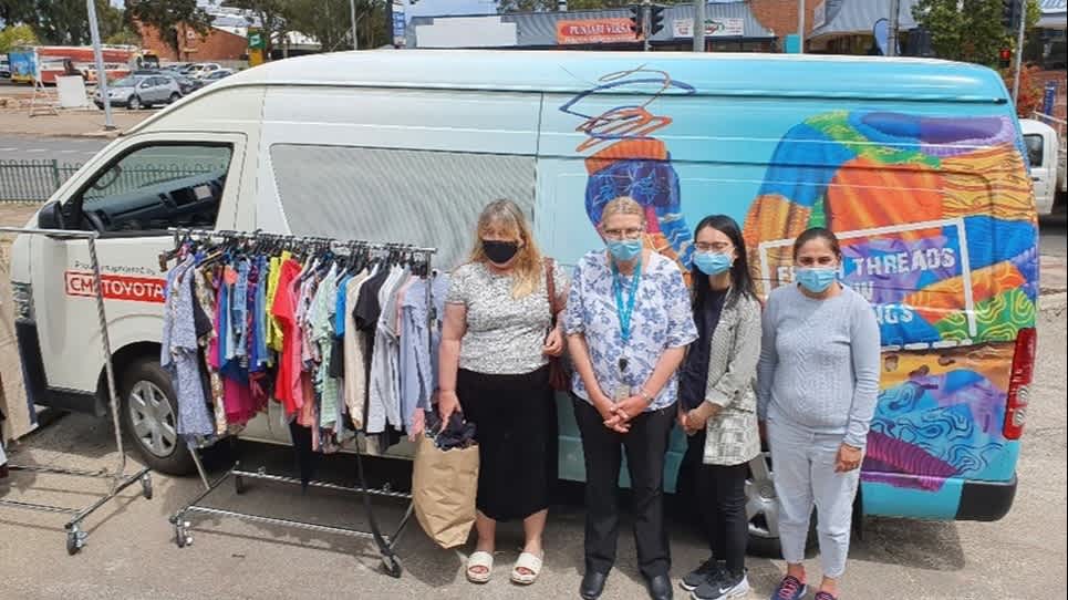 Four women standing infront of a van that has a clothes rack out the front.